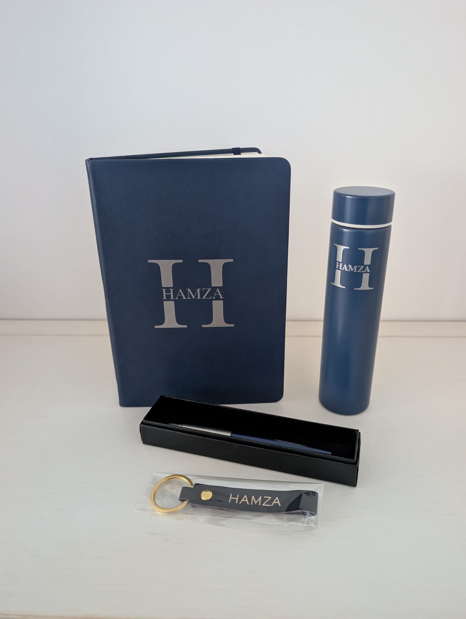 The four monogrammed items standing together on a pale wooden surface: navy notebook, navy insulated flask, pen in an open black presentation box and navy leather keyring with a gold ring, each with the same silver H monogram and the name HAMZA