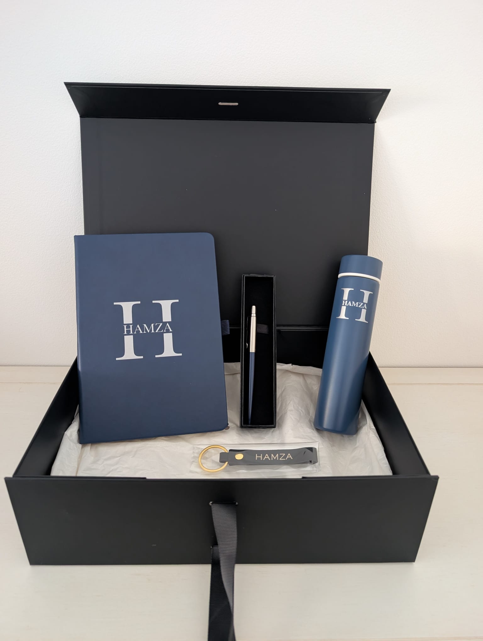 Open black magnetic gift box on white tissue holding a navy notebook, a boxed pen, a navy insulated flask and a navy leather keyring with a gold ring, all carrying the same silver H monogram with the name HAMZA