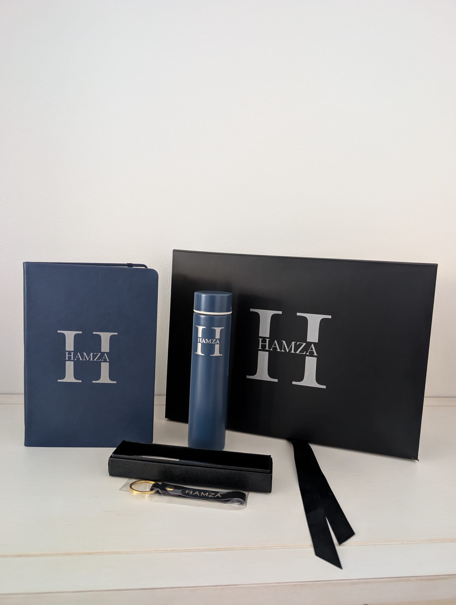 Navy monogrammed notebook, insulated flask, boxed pen and keyring in its clear packaging, standing in front of the closed black box lid with the same silver H monogram and a trailing black ribbon